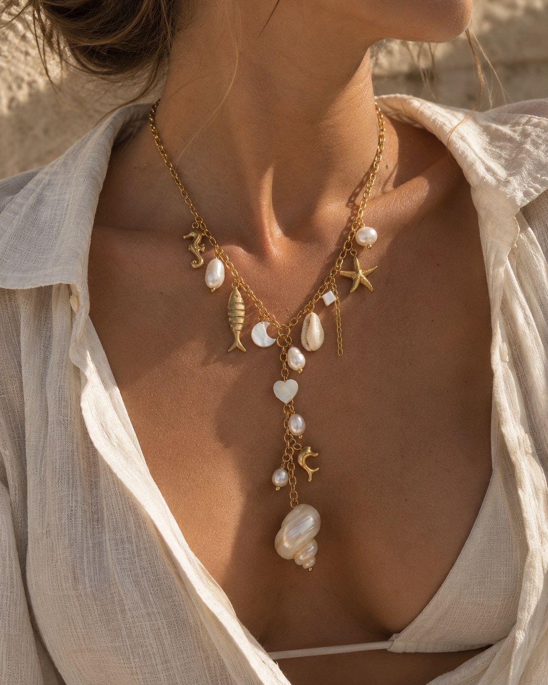 A close-up of a woman wearing a gold-toned layered necklace with white pearl-like beads, starfish, seashell, and fish charms, styled with an open white linen shirt.