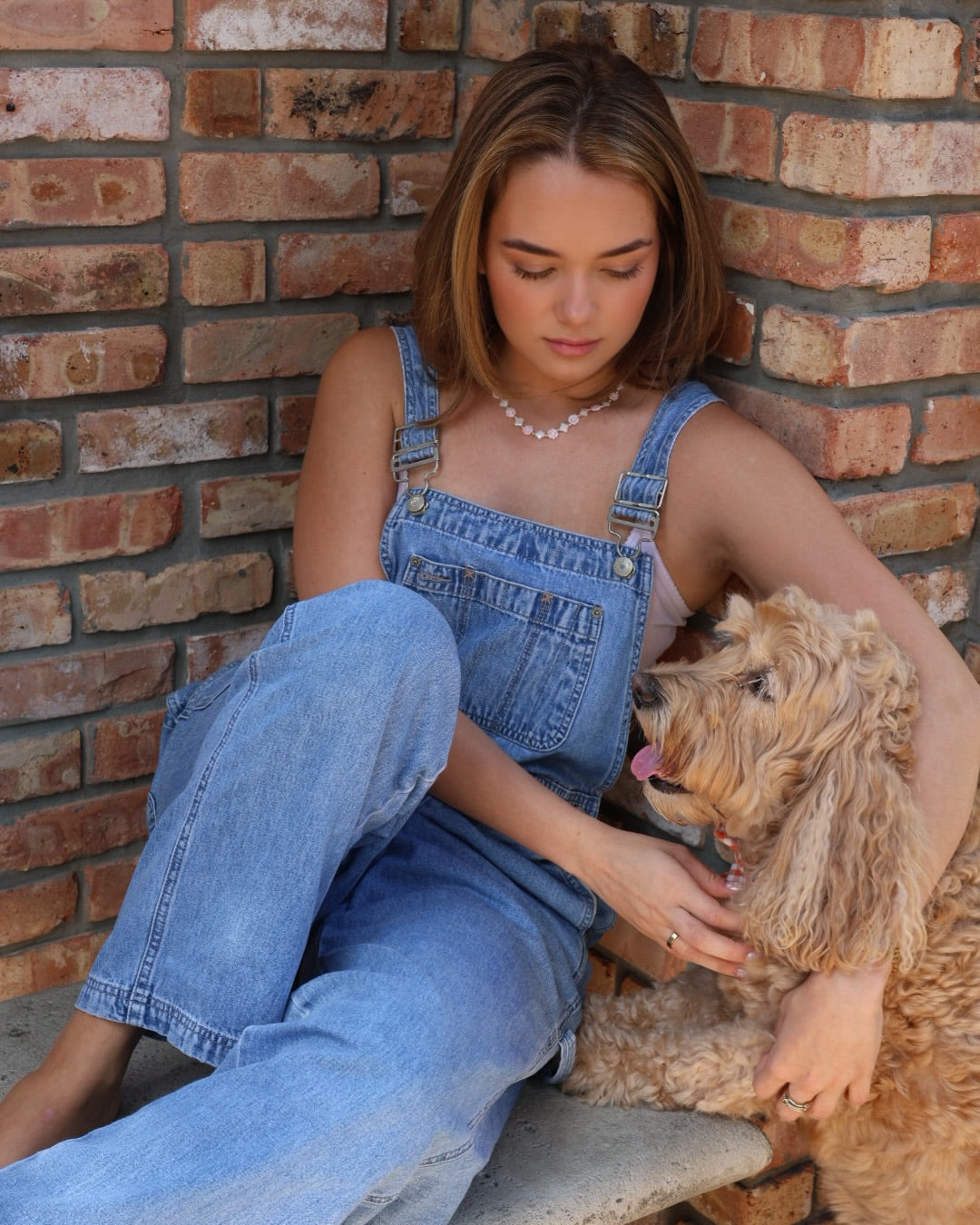 Woman in denim overalls sitting on a bench with a dog against a brick wall.