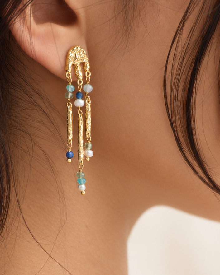 A close-up lifestyle shot of a model wearing a long, intricate gold dangle earring with small blue, white, and dark blue beads, paired with a delicate gold chain necklace and a white linen top.