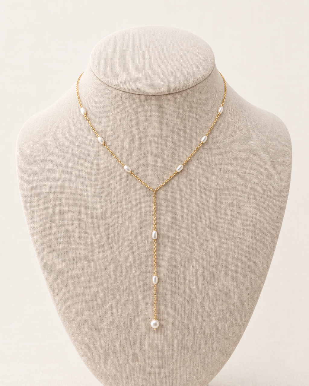 A delicate gold Y-necklace with small white pearls, displayed on a beige fabric jewelry bust against a light beige background.