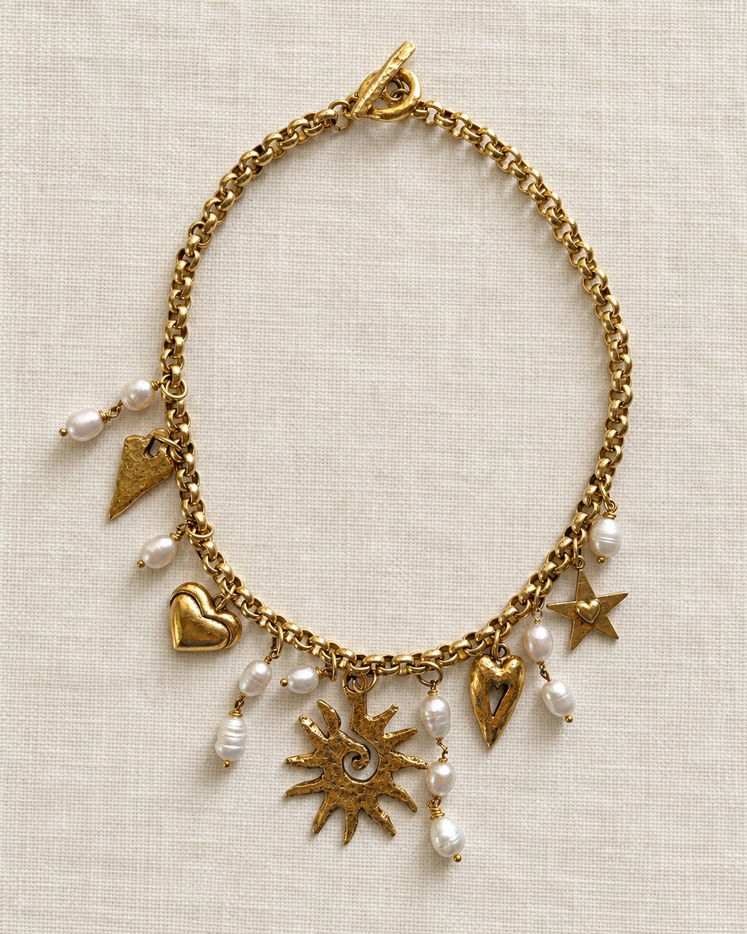 A gold-tone chain necklace with multiple charms, including hearts, stars, a sunburst, and small white pearl beads, displayed on a textured beige linen background.