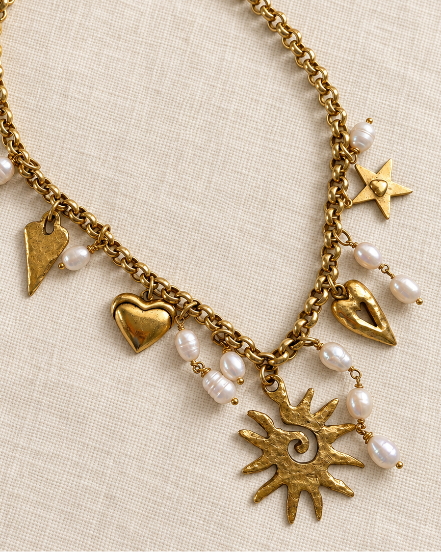 A gold-tone chain bracelet with heart, star, and sunburst charms, accented with small white pearl beads, displayed on a textured beige linen background.