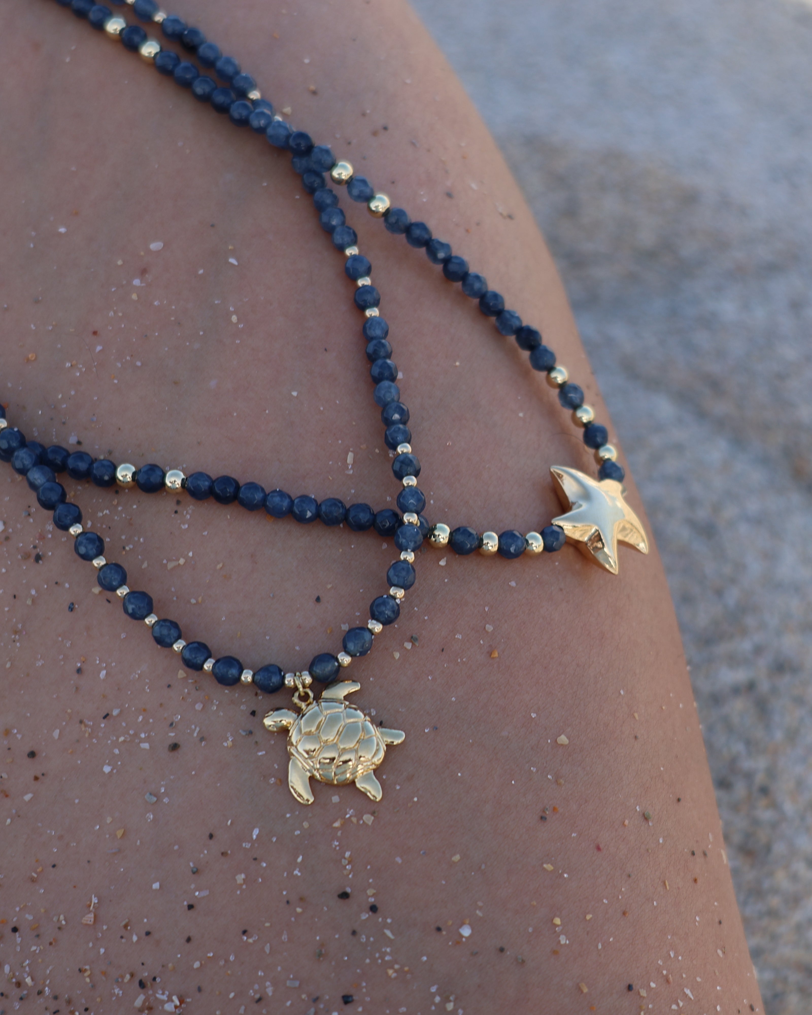 Navy beaded necklace with gold turtle pendant on a person's arm against a sandy background