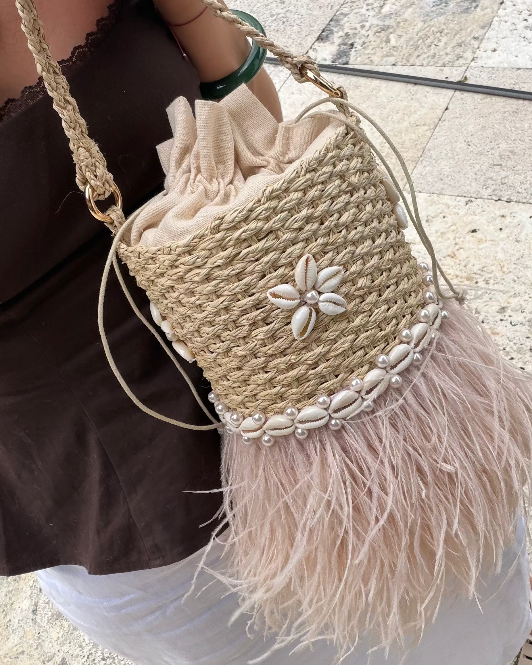 A woman holds a woven straw bucket bag with a beige drawstring top, a white shell flower detail, a band of white cowrie shells, and a light pink feather fringe. The bag has a braided shoulder strap and is shown in an outdoor setting on a stone floor.