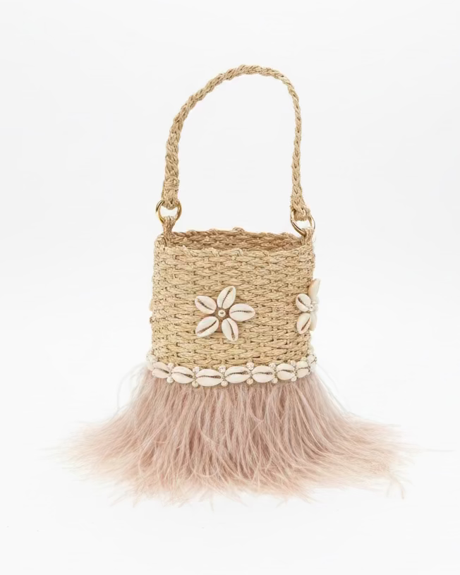 A woven straw handbag with a light pink feather skirt and white seashell trim, displayed on a gray slatted outdoor table.