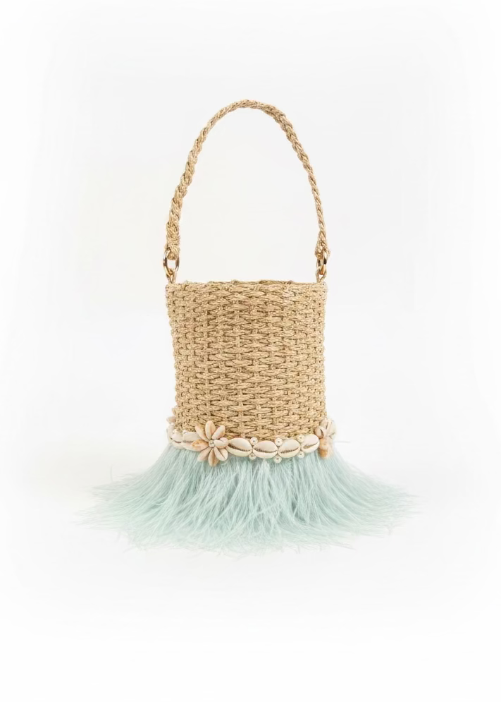 A natural straw bucket bag with a braided handle, decorated with a band of seashells and a light blue feather fringe, sitting on a dark gray slatted table.