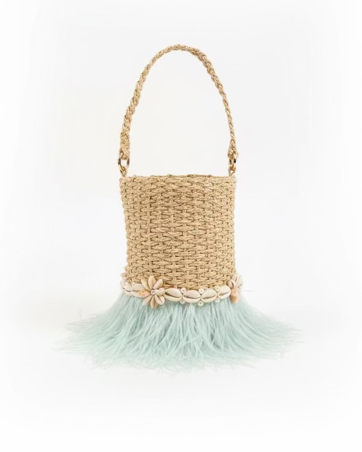 A natural straw bucket bag with a braided handle, decorated with a band of seashells and a light blue feather fringe, sitting on a dark gray slatted table.
