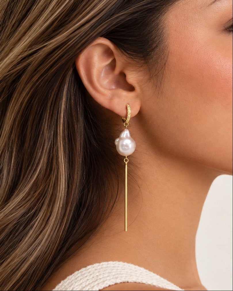 A woman in profile wearing a delicate gold dangle earring with a white freshwater pearl, shown against a clean off-white background.