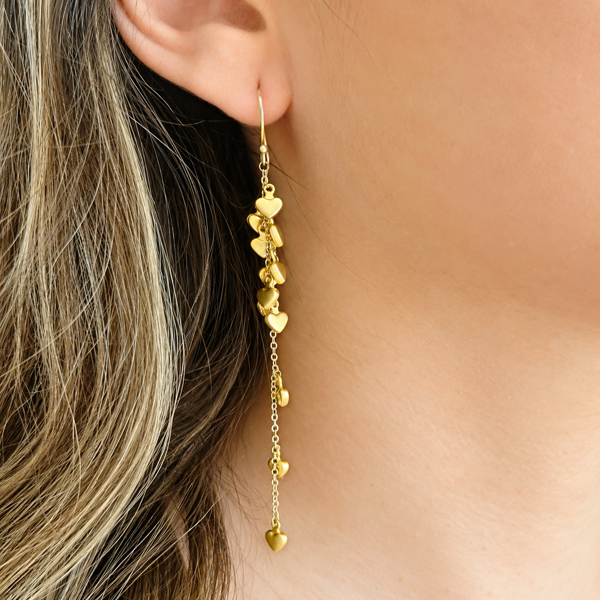 A close-up of a gold dangle earring with multiple small heart charms, shown worn on a person's ear.