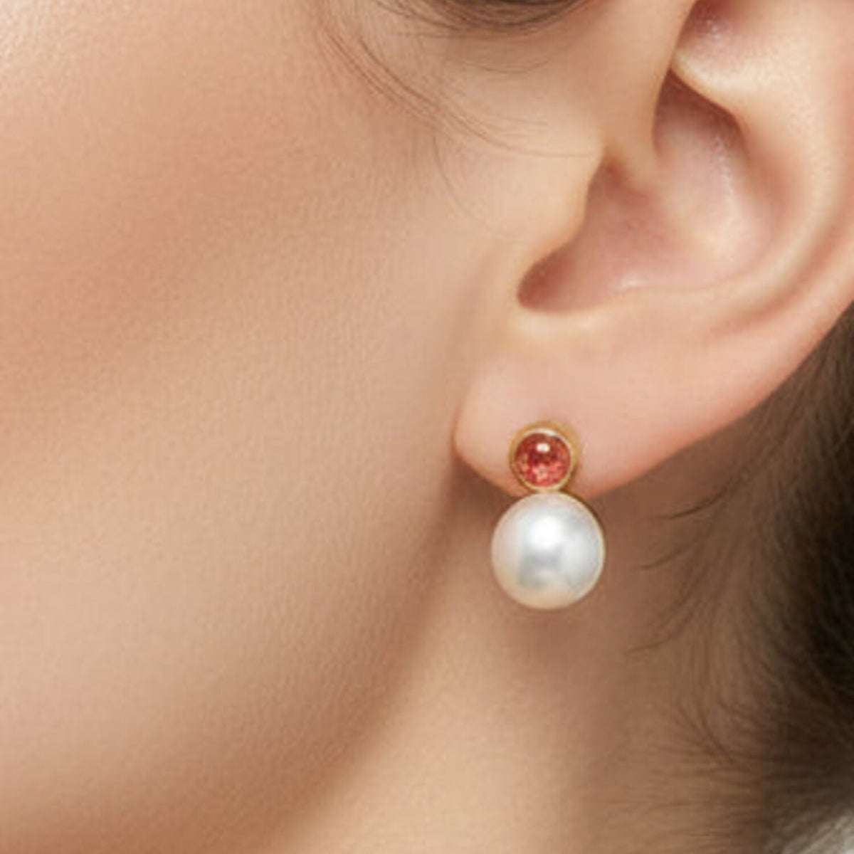 A close-up of a model's ear wearing a gold stud earring with a small round red gemstone and a dangling white pearl.