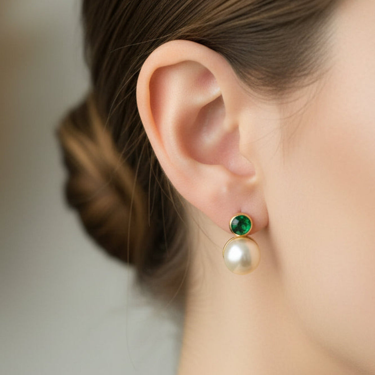 A close-up of a model's ear wearing a drop earring with a small round green gemstone and a larger white pearl below, set in gold-toned metal.