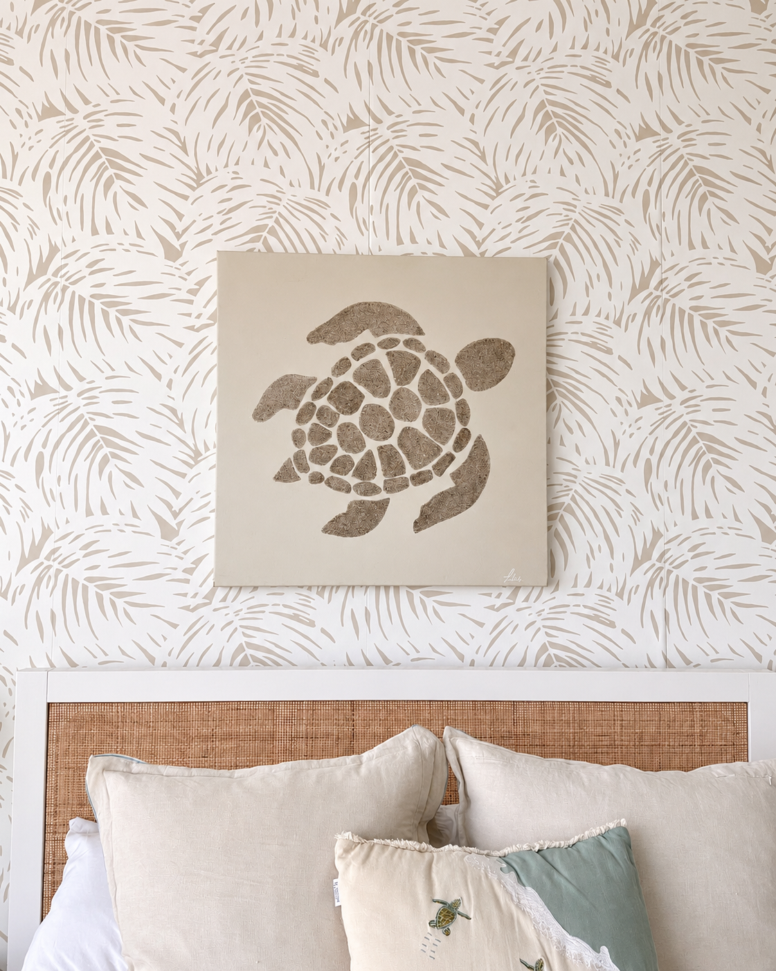 A square canvas print featuring a brown sea turtle silhouette on a cream background, displayed on a wall above a bed with neutral-toned pillows and a woven headboard.