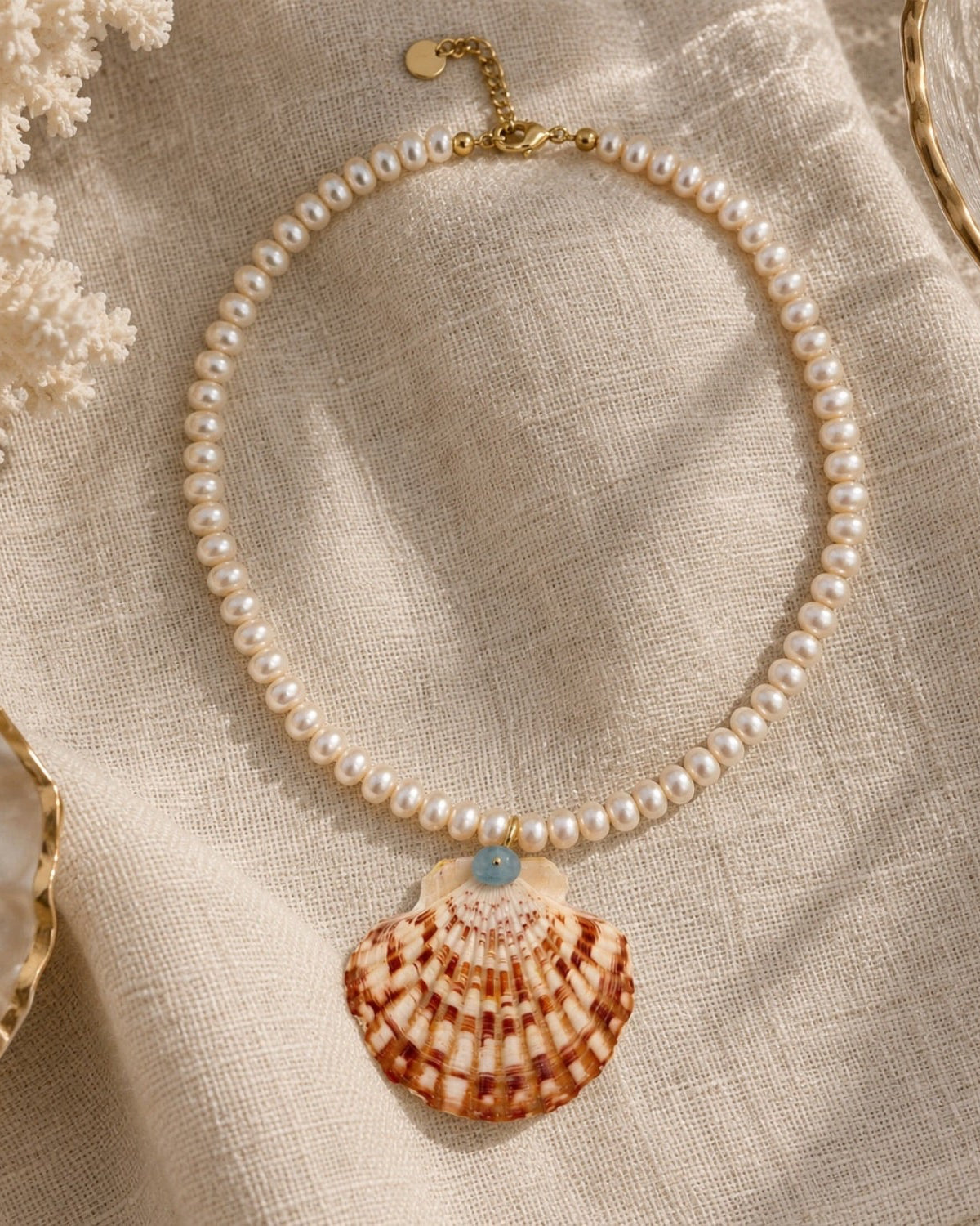 A pearl necklace with a large seashell pendant and a small light blue bead, styled on a beige linen fabric with coral and shell props.