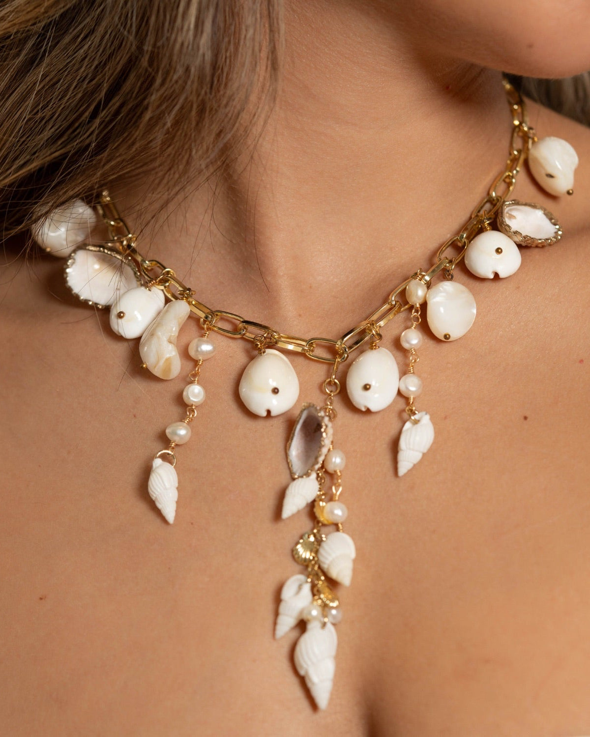 A close-up of a woman wearing a gold-toned statement necklace adorned with white shell beads, pearl-like beads, and dangling leaf-shaped charms.