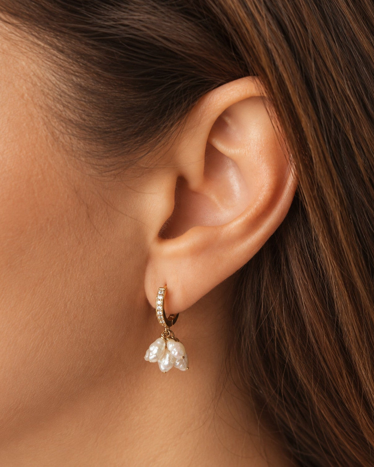A close-up of a model's ear wearing a small gold huggie hoop earring with a dangling cluster of white faux pearls.