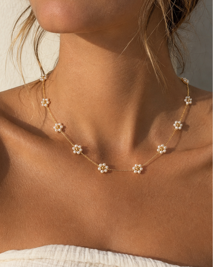 A close-up of a model wearing a delicate gold chain necklace adorned with small white daisy flowers, styled with a white textured top against a neutral background.