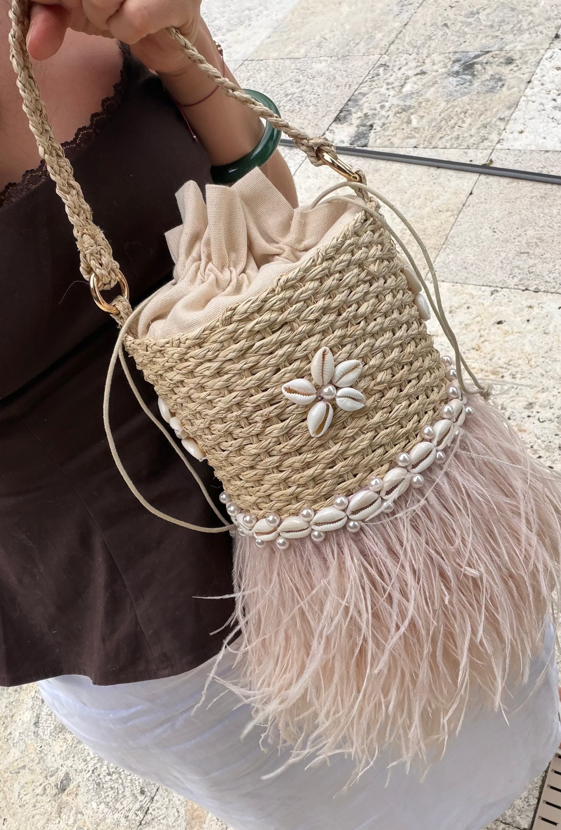 A woman holds a woven straw bucket bag with a beige drawstring top, a white shell flower detail, a band of white cowrie shells, and a light pink feather fringe. The bag has a braided shoulder strap and is shown in an outdoor setting on a stone floor.