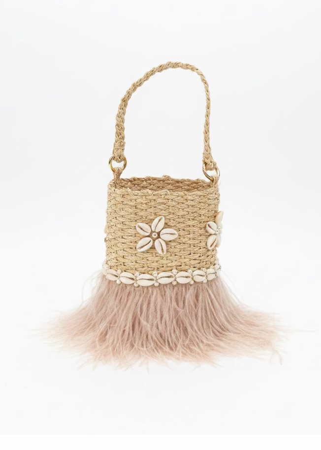 A woven straw handbag with a light pink feather skirt and white seashell trim, displayed on a gray slatted outdoor table.