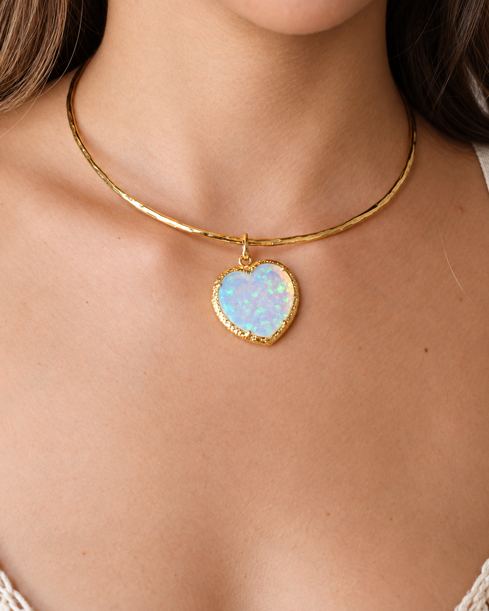 A close-up of a model wearing a gold bangle necklace with a heart-shaped iridescent opal pendant.
