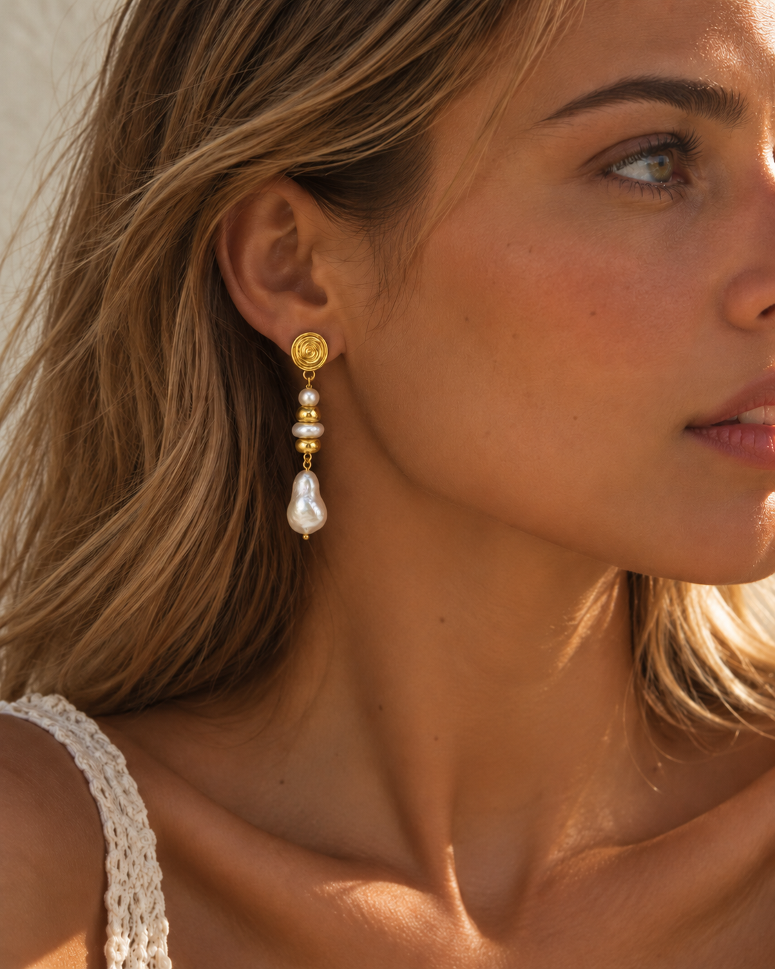 A close-up of a woman wearing a gold and pearl drop earring, showing the side profile of her head and ear against a warm, sunlit background.
