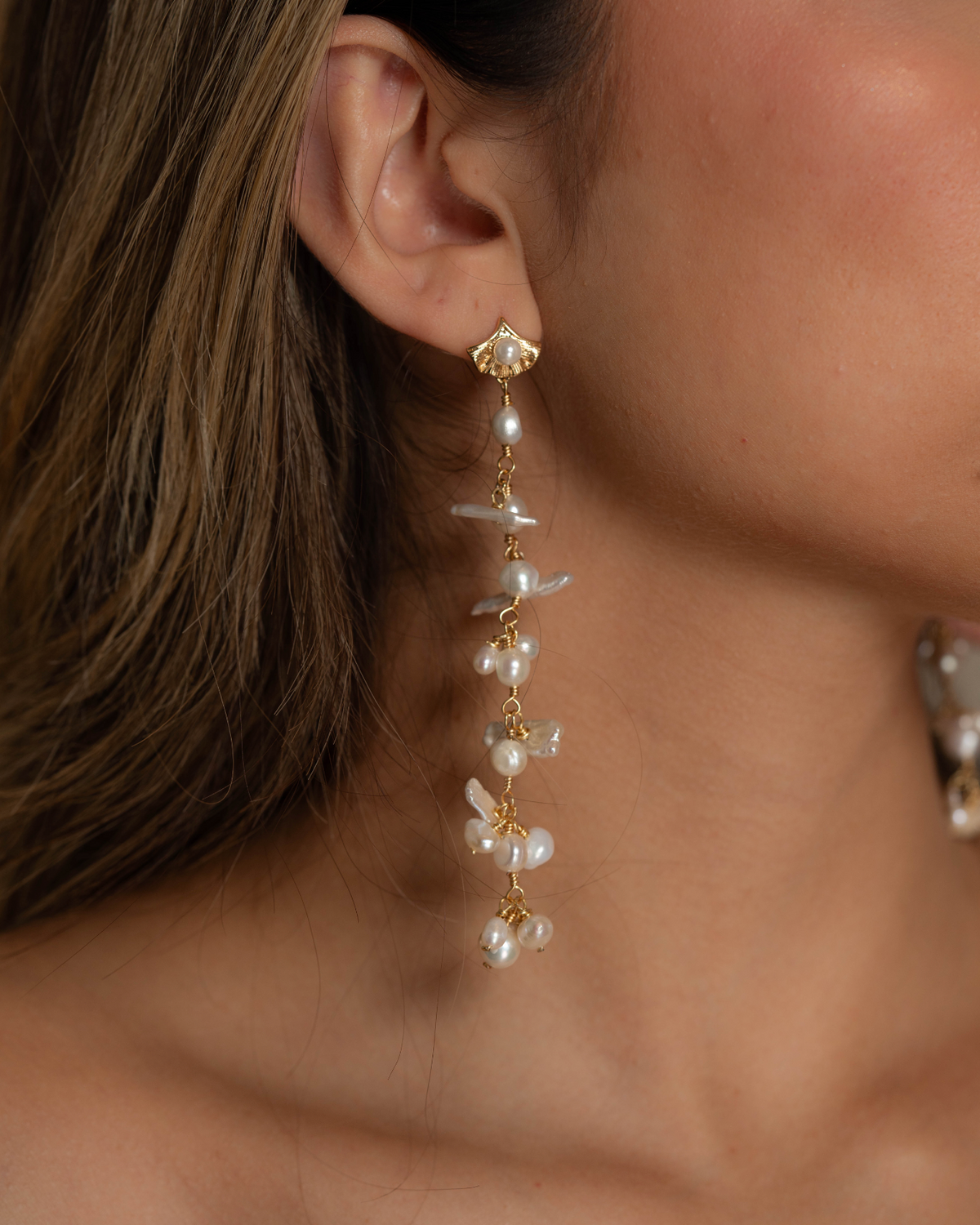 A close-up image of a woman wearing a long, gold-colored pearl earring with intricate designs.