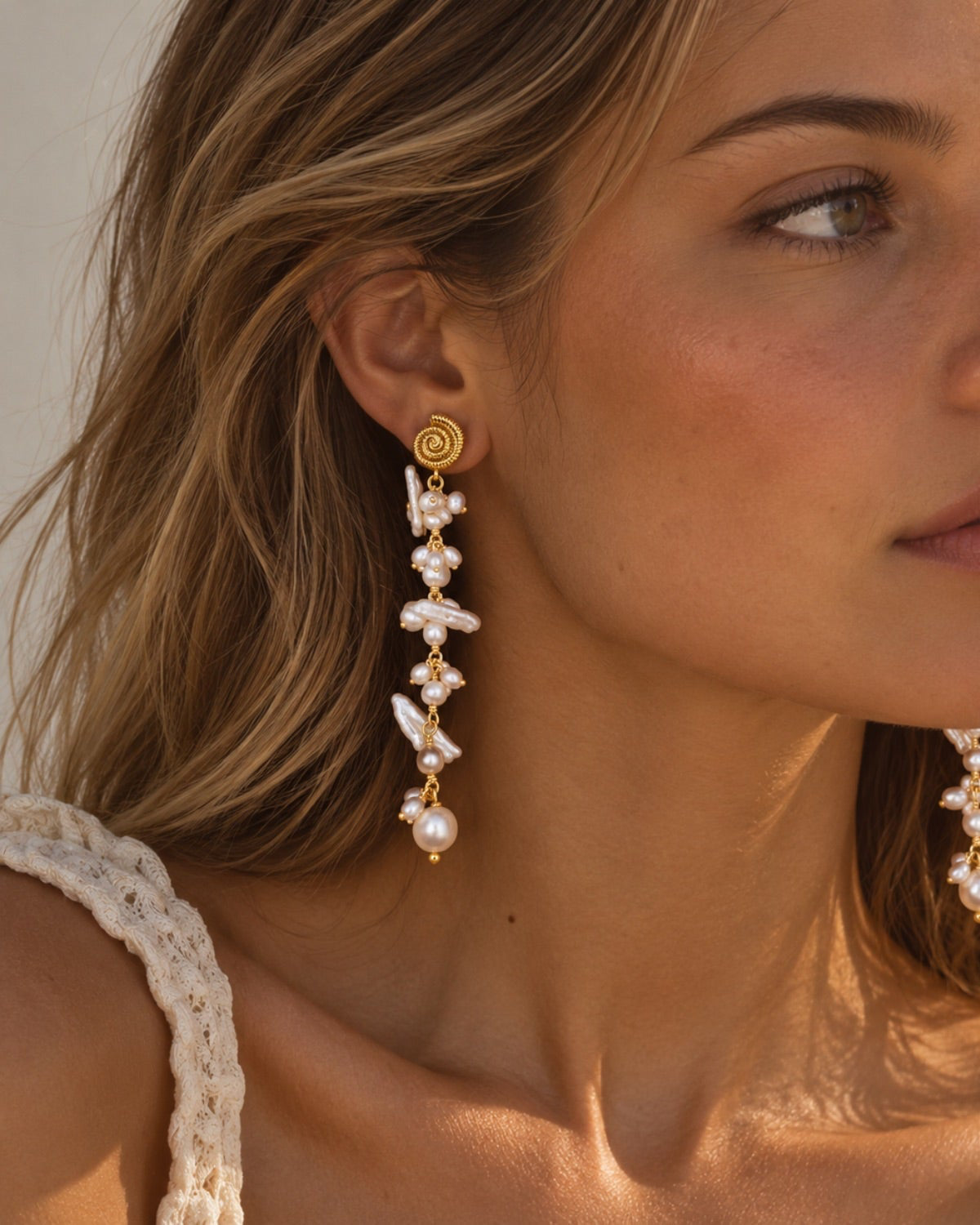 A close-up of a woman wearing a long, dangling statement earring with irregular white pearl-like beads and gold-toned metal details, including a spiral stud.