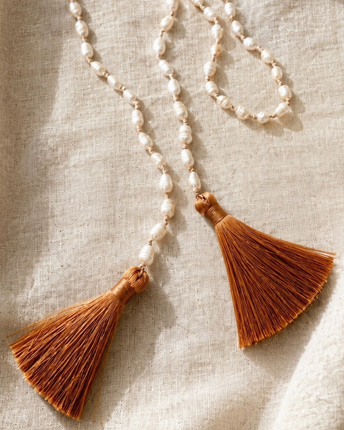 A bohemian-style beaded necklace with white pearl-like beads and two large brown tassel charms, laid out on a textured beige linen fabric.