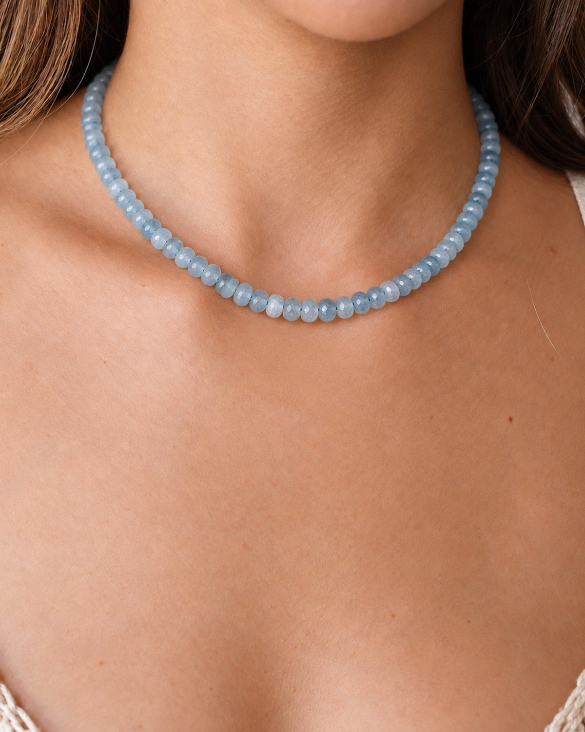 A close-up of a woman wearing a light blue beaded necklace with a matching light blue stone face pendant, paired with a cream crochet top.