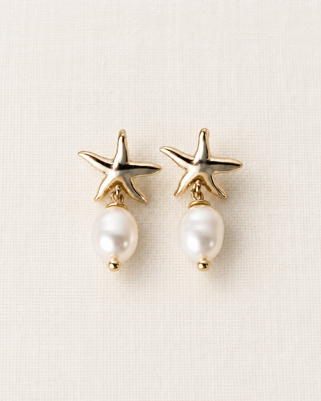 A pair of gold starfish stud earrings with dangling white pearl drops, displayed on a textured off-white fabric background.